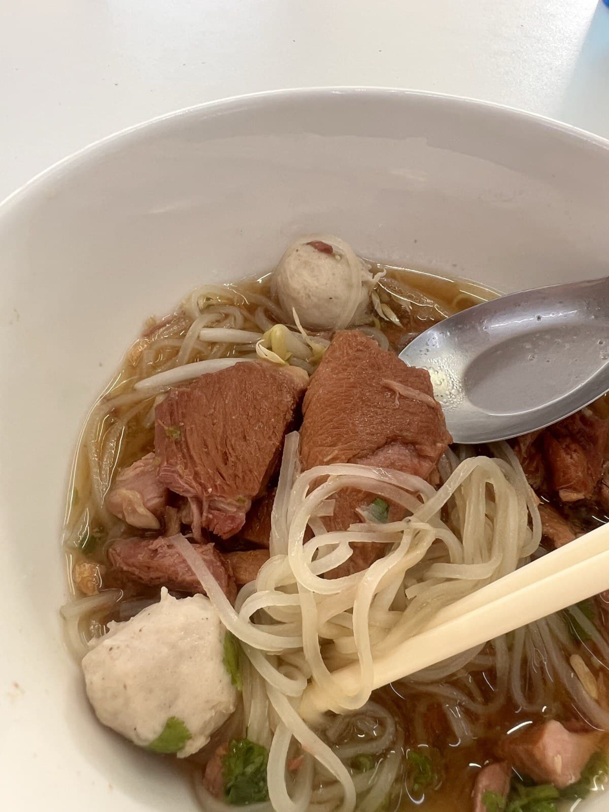 Chopsticks lifting thin rice noodles beside chunks of tender braised pork and two pork balls, a spoon resting in the broth